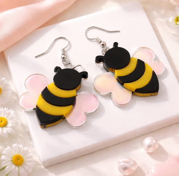 Bee-shaped acrylic dangle earrings with black and yellow bodies and iridescent wings on a soft neutral background